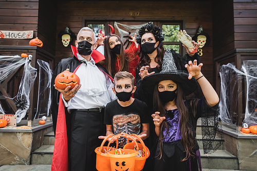 family in halloween costumes and black medical masks standing with buckets of sweets near house