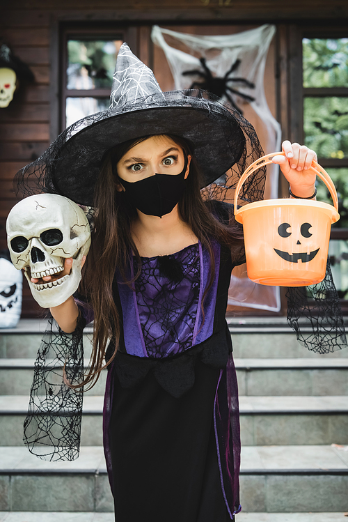 girl in witch halloween costume and black medical mask holding skull and bucket near blurred house