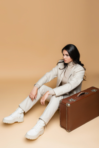 young woman in suit sitting near vintage suitcase on beige