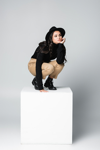 full length of stylish model in fedora hat looking away while sitting on white cube on grey
