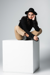 full length of stylish model in fedora hat  while sitting on white cube on grey