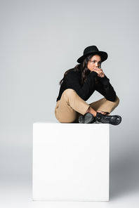 full length of pensive model in fedora hat sitting with crossed legs on white cube on grey