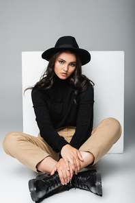 full length of stylish model in fedora hat sitting with crossed legs near white cube on grey
