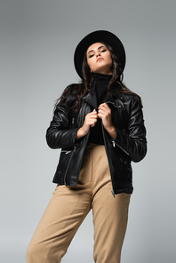 low angle view of trendy woman in fedora hat and black leather jacket  while posing isolated on grey