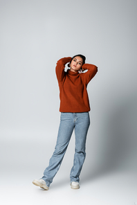 full length of young woman in sweater and denim jeans posing on grey