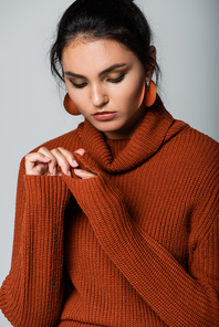 young woman in earrings and knitted sweater looking down isolated on grey