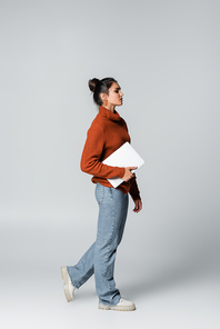 full length of young woman in knitted sweater and jeans holding laptop on grey