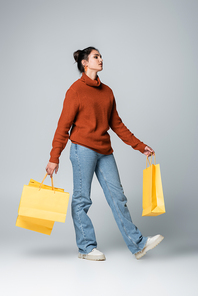 full length of young model in sweater and jeans holding yellow shopping bags on grey