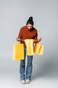 full length of young woman in sweater and jeans holding yellow shopping bags on grey