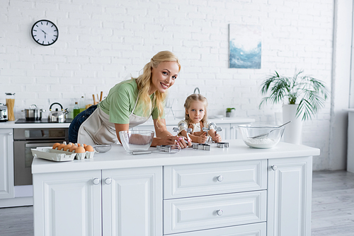 woman smiling at camera near daughter and table with ingredients in kitchen