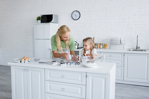 mother holding bowl near daughter and kitchen table with ingredients