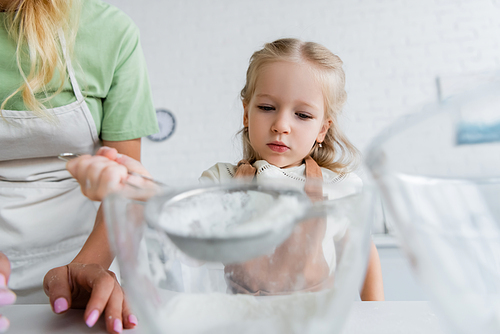 child sifting flour into bowl near mom in kitchen, blurred foreground