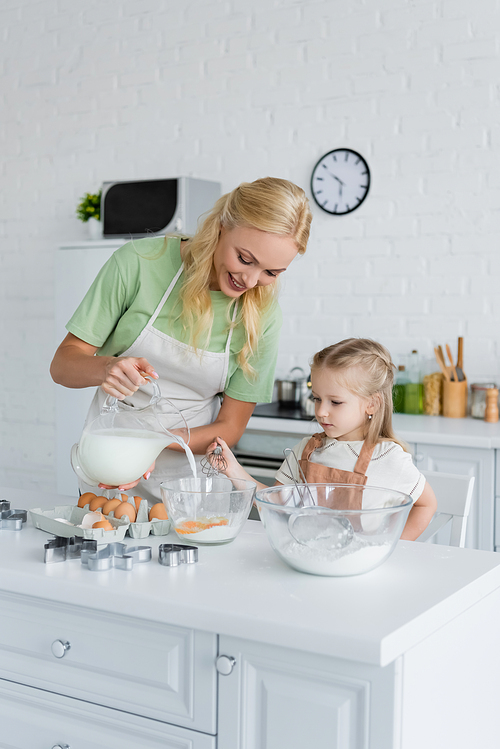 smiling woman pouring milk into bowl near daughter with whisk