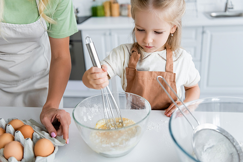 girl in apron mixing chicken eggs with flour in bowl near mom in kitchen