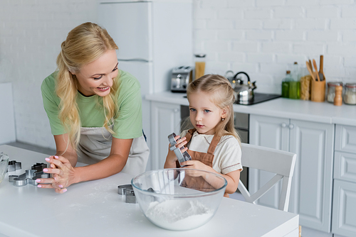 child in apron holding cookie cutter near happy mom and bowl with flour