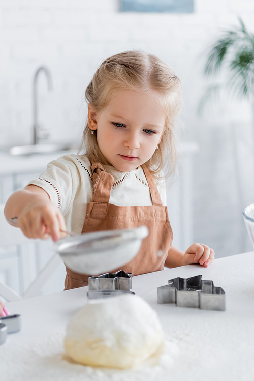 child in apron sifting flour near cookie cutters, blurred foreground