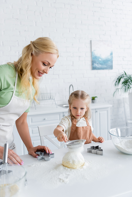 child sifting flour on raw dough while helping mom in kitchen