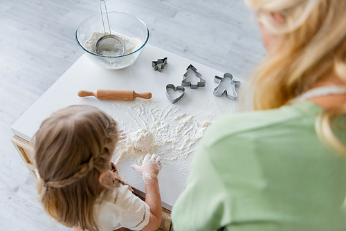 overhead view of girl kneading dough near blurred mom in kitchen