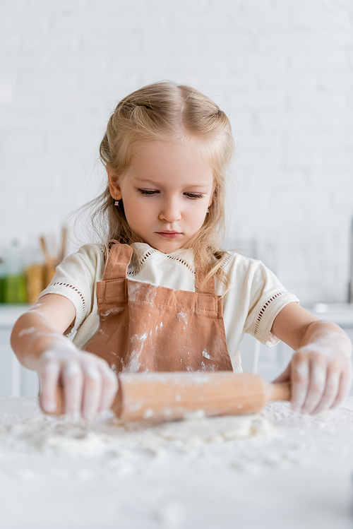 kid in apron rolling dough in kitchen on blurred foreground