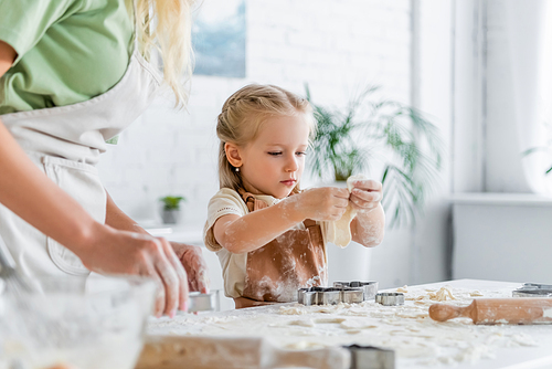 little girl holding dough near blurred mother and cookie cutters on kitchen table