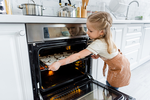 girl in apron placing baking sheet with cookies in oven