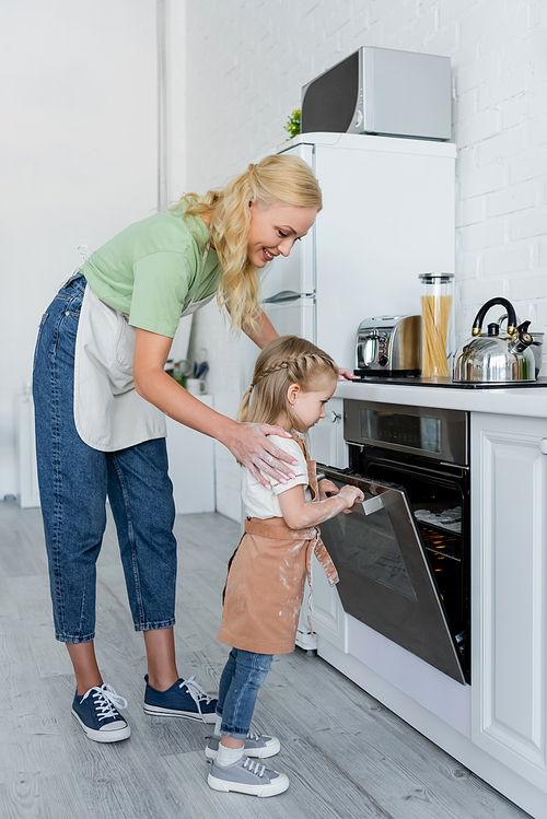 girl looking at cookies in electric oven near happy mother