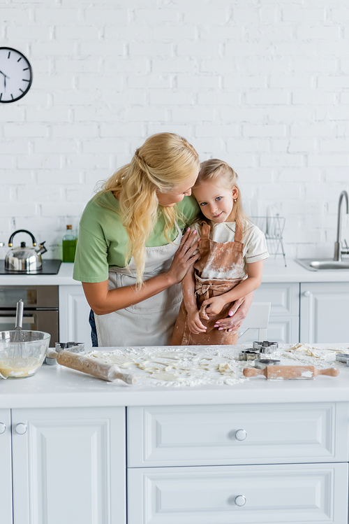 woman hugging daughter near kitchen table with flour and cooking utensils