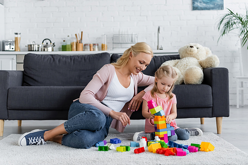 girl making tower of building blocks near mom and teddy bear on couch