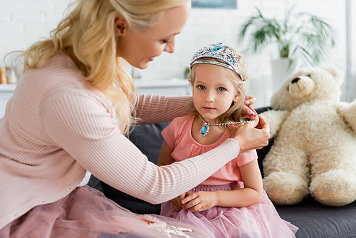 blurred woman putting toy necklace on daughter while having fun at home