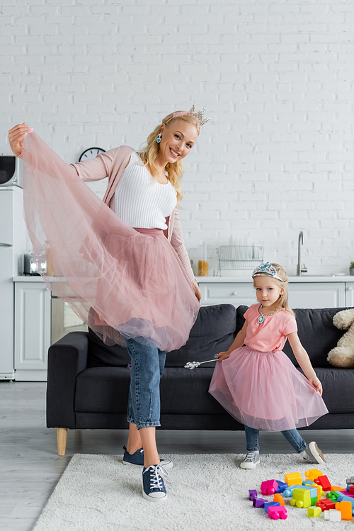 cheerful mom and daughter in toy crowns and tulle skirts  while dancing at home