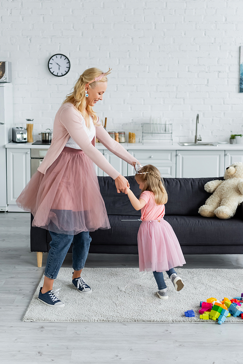 happy woman and daughter in costumes of princesses dancing in kitchen