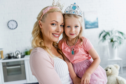 happy mom and daughter in toy crowns smiling at camera at home