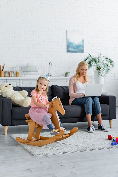 child in toy crown riding rocking horse near blurred mom working on laptop