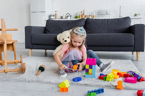 girl in toy crown playing with magic wand and colorful cubes on floor near sofa and teddy bear