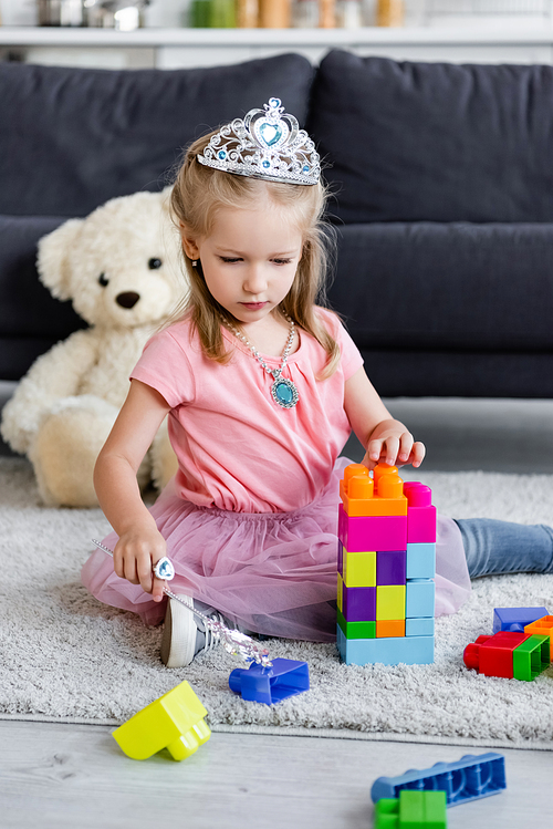 girl in costume of princess playing with magic wand and building blocks near blurred teddy bear