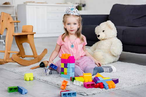 child in toy crown and necklace  near building blocks and blurred teddy bear