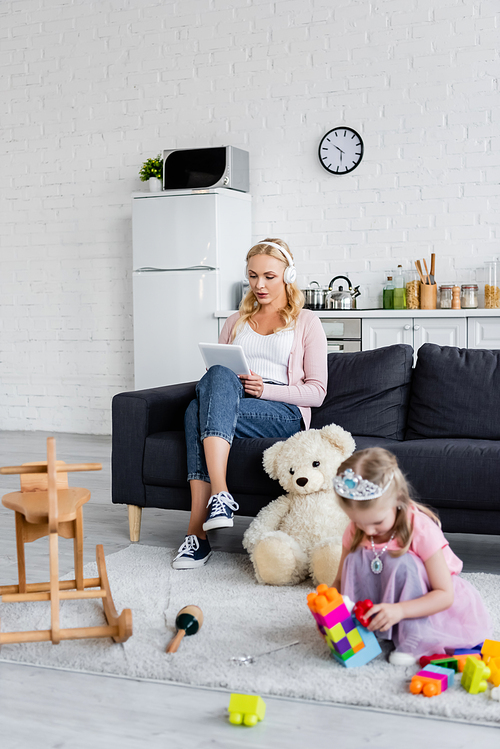 blurred girl playing on floor near mom with digital tablet listening music in headphones