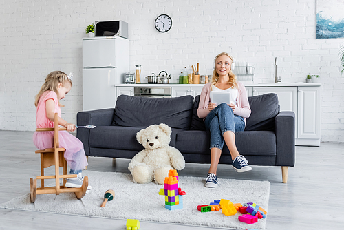 happy woman listening music on sofa near daughter in toy crown riding rocking horse