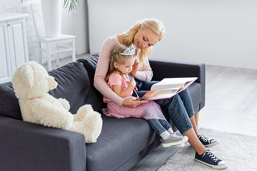 smiling mother touching hair of girl in toy crown while sitting on sofa with book