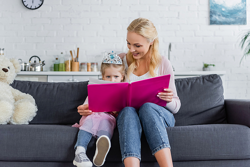 mom reading book to daughter in toy crown while sitting on sofa at home