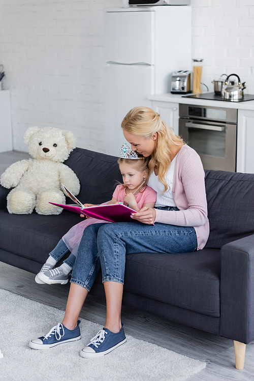 mother reading book to daughter in toy crown near teddy bear on couch