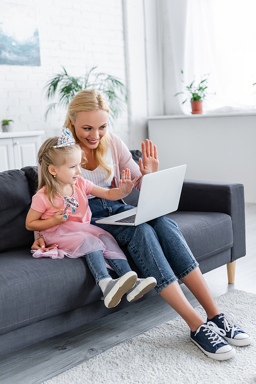 cheerful woman and girl in toy crown waving hands during video call on laptop