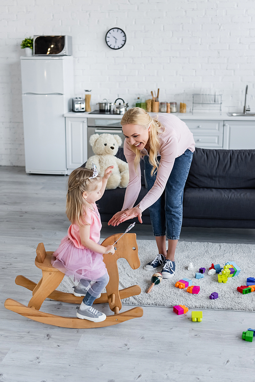 happy woman giving hands to daughter riding rocking horse in costume of princess