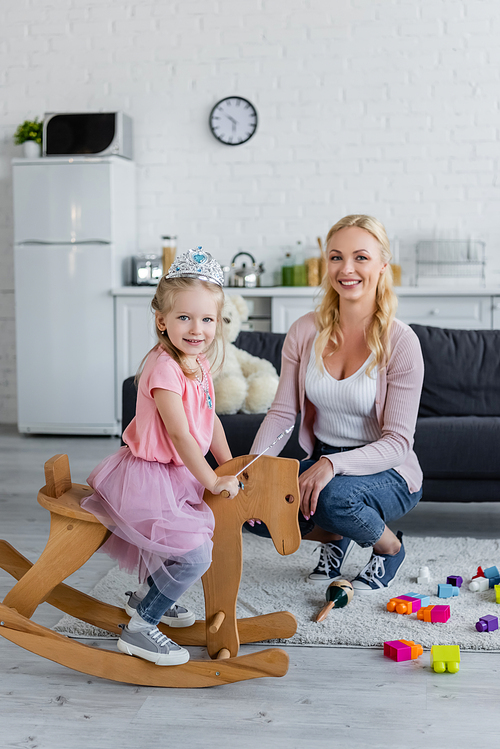 happy child in fairy costume riding rocking horse near smiling mother