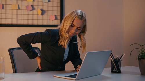 Businesswoman suffering from back pain near laptop in office in evening