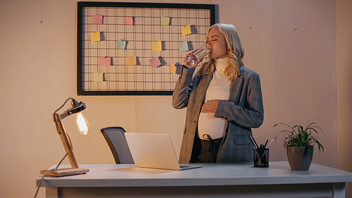 Pregnant businesswoman drinking water near laptop in office