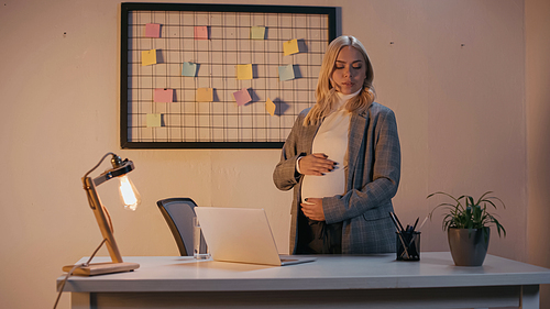 Pregnant businesswoman standing near stationery and laptop in office in evening