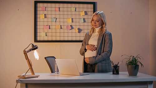 Pregnant businesswoman touching belly near laptop and glass of water in office in evening