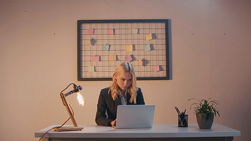 Businesswoman using laptop near lamp and stationery in office in evening