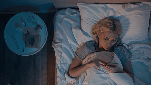 Top view of young woman using smartphone near pills and electronic thermometer on bed